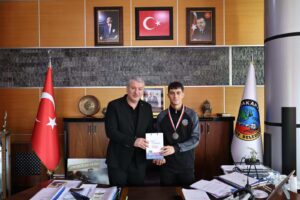 Alt metin: Belediye başkanı ve genç sporcu, ödül takdiminde resmi ortamda, Türk bayrağı ve resmi portrelerle çevrili.