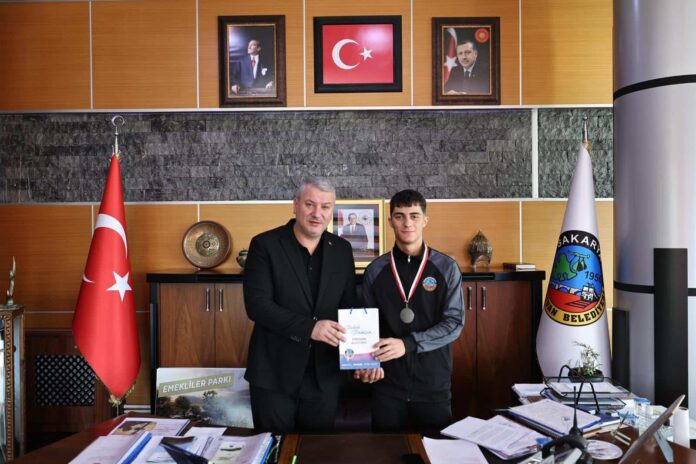 Alt metin: Belediye başkanı ve genç sporcu, ödül takdiminde resmi ortamda, Türk bayrağı ve resmi portrelerle çevrili.